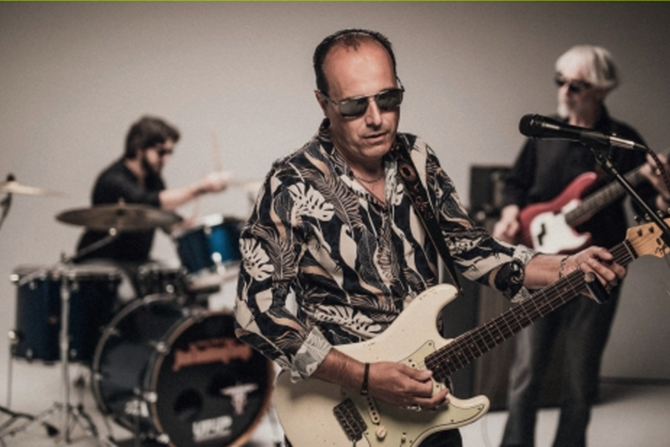 Eine Band spielt live auf der Bühne. Der Sänger trägt eine Sonnenbrille und spielt eine E-Gitarre, während die anderen Musiker am Schlagzeug und Bass stehen.