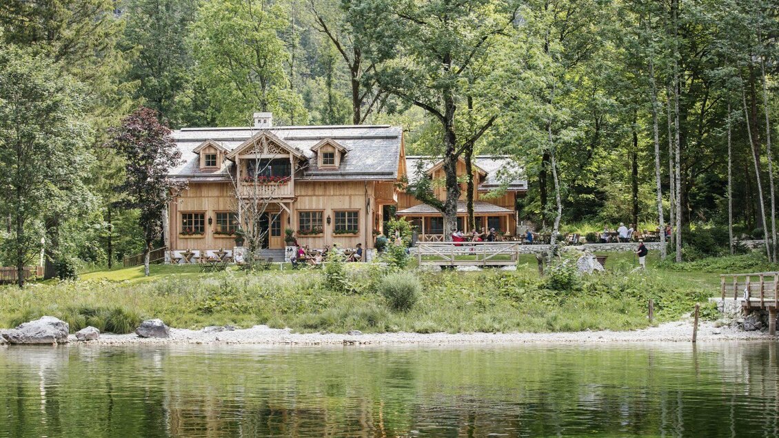 Eine Außenansicht der TAUROA Seewiese aus Richtung des Altausseer Sees. Es ist ein traditionelles, charmantes Holzhaus am Ufer des Altausseer Sees, umgeben von grünen Bäumen und der spektakulären Landschaft des Naturplatzes Seewiese in Altaussee. | © Tauroa