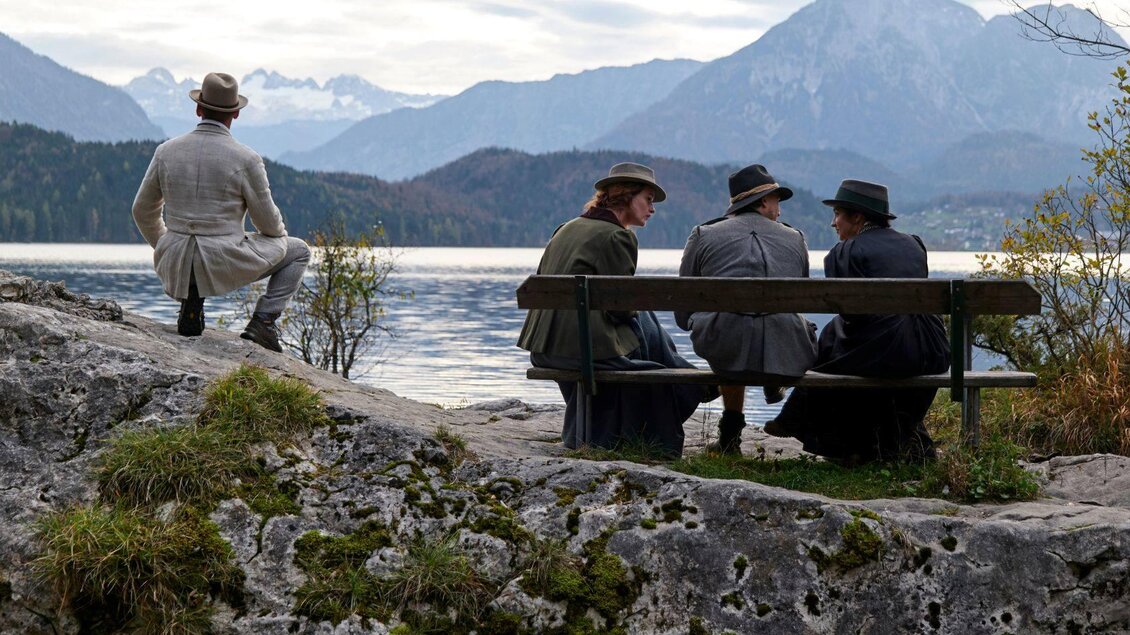 Eine Gruppe von vier Personen in historischer Kleidung sitzt auf einer Bank am Ufer eines Sees. Im Hintergrund sind Berge und ein bewölkter Himmel zu sehen. | © Tauroa