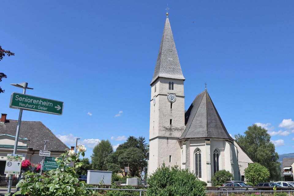 Eine alte Kirche mit einem hohen Turm steht im Zentrum des Bildes. Im Vordergrund sind grüne Pflanzen und ein Wegweiser zu sehen. | © Anita Fössl