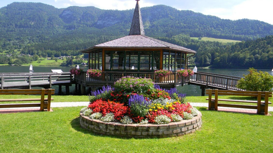 Ein schöner Pavillon am See, umgeben von bunten Blumenbeeten. Im Hintergrund sind grüne Berge und ein blauer Himmel zu sehen. | © TVB Ausseerland - Salzkammergut/Gigler