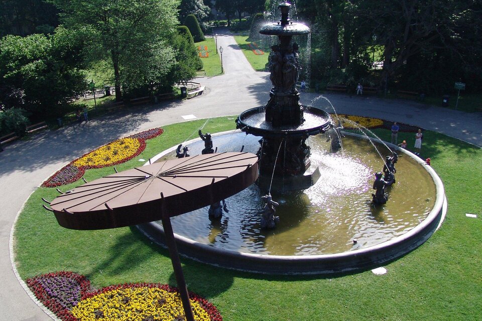 Ein schöner Brunnen in einem Park mit blühenden Blumenbeeten. Umgeben von Bäumen und einem gepflegten Weg.