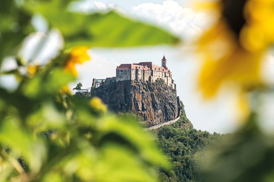 Eine beeindruckende Burg auf einem Felsen, umgeben von grüner Landschaft. Im Vordergrund sind farbenfrohe Sonnenblumen zu sehen. | © Rudi Ferder