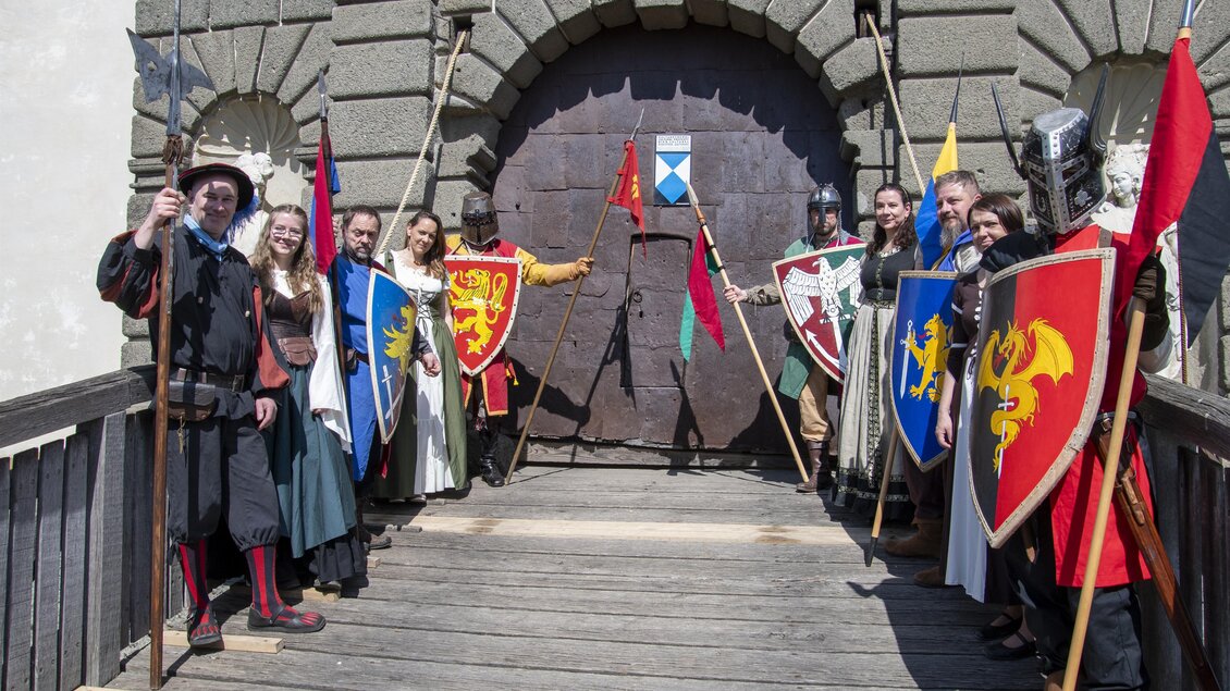 Eine Gruppe von Rittern und Burgbewohnern steht vor einem großen Holztor. Sie tragen historische Kostüme und halten Schilde mit verschiedenen Wappen. | © Tourismusverband Thermen- & Vulkanland/Christian Thomaser