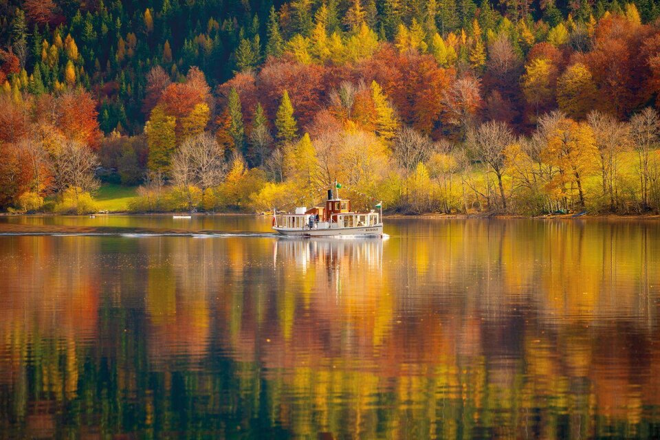 Ein nostalgisches Schiff fährt über einen ruhigen See, umgeben von bunten Bäumen im Herbst. Die leuchtenden Farben spiegeln sich im Wasser wider. | © Schifffahrt Grundlsee