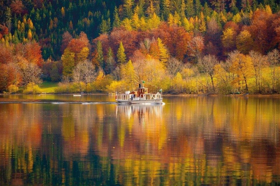 Die nostalgische MS Rudolf der Schifffahrt Grundlsee fährt ruhig über den klaren Grundlsee. Am Ufer erstrahlt die malerische Landschaft im bunten Herbstlaub. | © Schifffahrt Grundlsee