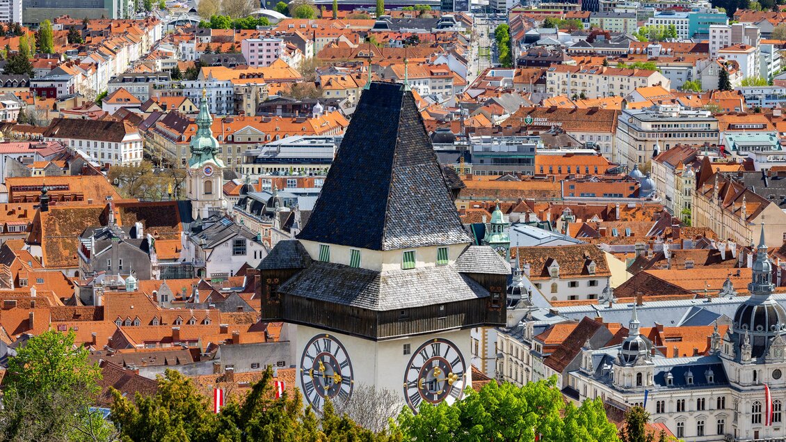 Eine Stadtansicht mit vielen roten Dächern und einem markanten Uhrturm im Vordergrund. Die Landschaft wird von grünen Bäumen umgeben und zeigt eine lebendige Stadt. | © Graz Tourismus - Harry Schiffe