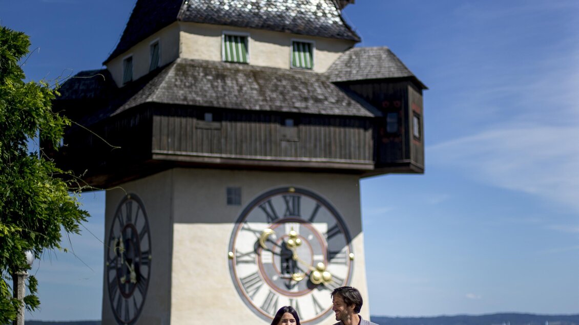 Ein Paar spaziert vor einem historischen Uhrturm mit einem klaren blauen Himmel im Hintergrund. Die Umgebung ist grün und gepflegt, was eine angenehme Atmosphäre schafft. | © (c) Graz Tourismus - Tom Lamm