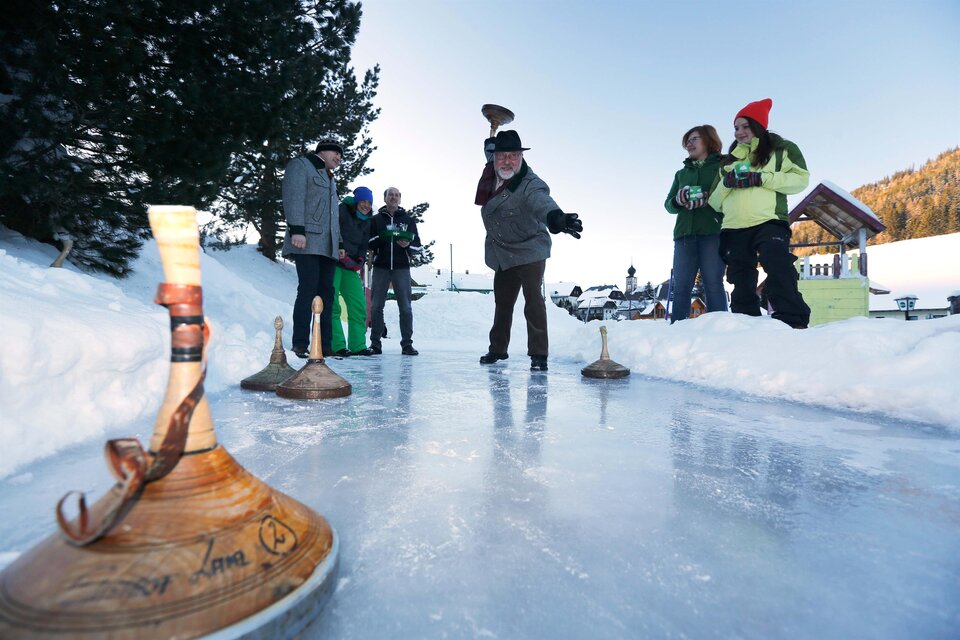 Eine Gruppe von Menschen spielt auf einem gefrorenen Teich im Schnee. Es sind verschiedene Holzgeräte zu sehen, die für das Spiel verwendet werden. | © Herbert Raffalt