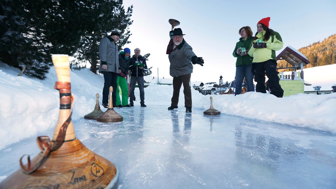 Eine Gruppe von Menschen spielt auf einem gefrorenen Teich im Schnee. Es sind verschiedene Holzgeräte zu sehen, die für das Spiel verwendet werden. | © Herbert Raffalt