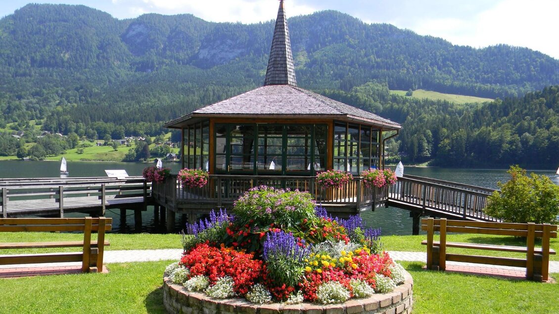 Ein schöner Pavillon am See, umgeben von bunten Blumen. Im Hintergrund sind grüne Hügel und ein klarer Himmel zu sehen. | © Gemeinde Grundlsee