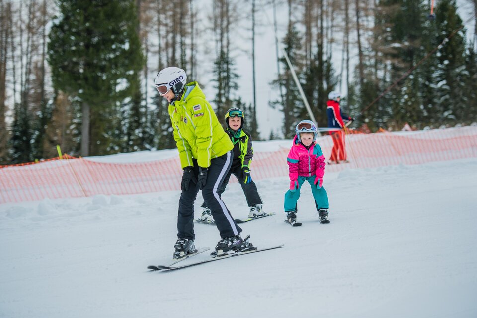 Eine Skilehrerin mit zwei Kindern fährt auf einer schneebedeckten Piste. Im Hintergrund sind Bäume und eine orangefarbene Absperrung zu sehen. | © Tourismusverband Murtal