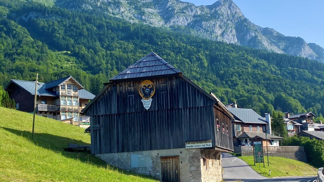 Ein traditionelles Holzhaus mit einem Wappen, umgeben von grünen Wiesen und Bergen. Im Hintergrund sind weitere Gebäude und eine klare blaue Himmel zu sehen. | © Bettina Scheck