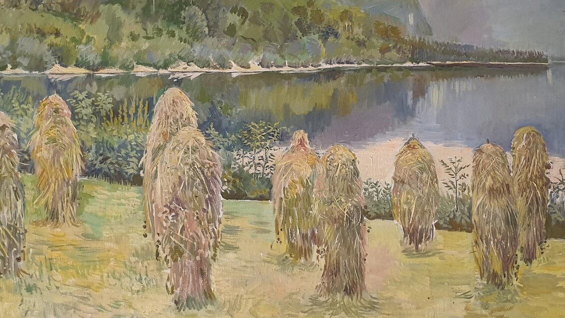 Eine friedliche Landschaft mit einem See und überhängenden Bäumen im Hintergrund. Im Vordergrund stehen Strohfiguren auf einer Wiese. | © A. Schoiswohl