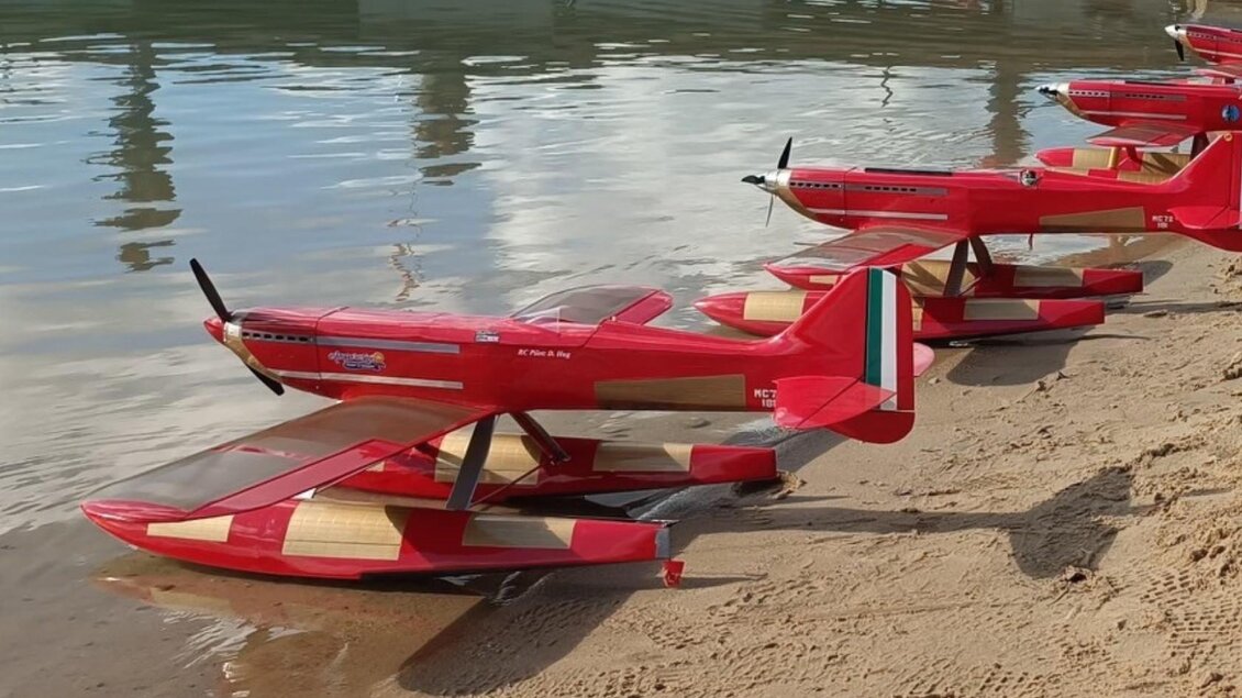 Eine Reihe roter Modellflugzeuge steht am Ufer eines Gewässers. Der Sandstrand und das ruhige Wasser schaffen eine entspannte Atmosphäre. | © Modell Wasserflugverein Lausitz e. V.