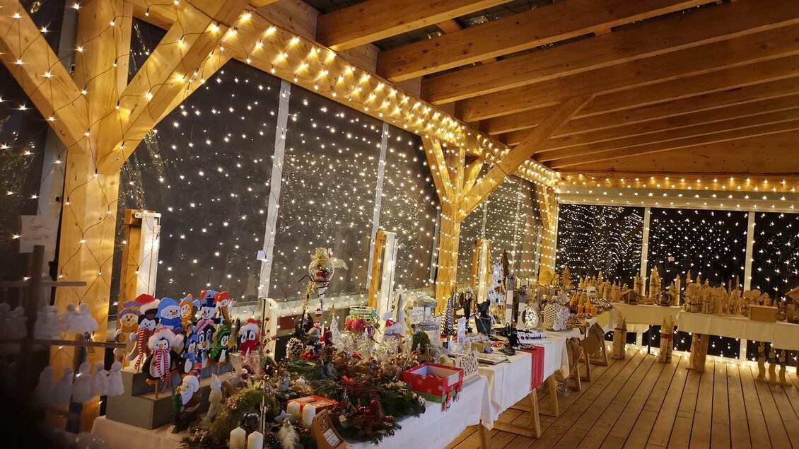 Ein festlich dekorierter Raum mit vielen Lichtern und einer Holzstruktur. Auf den Tischen sind verschiedene handgefertigte Weihnachtsartikel ausgestellt. | © Flourl´s Schenke