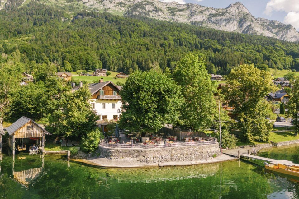 Eine idyllische Landschaft mit einem klaren See und umgeben von Bäumen. Im Hintergrund sind Berggipfel und traditionelle Häuser zu sehen. | © DraganTadic
