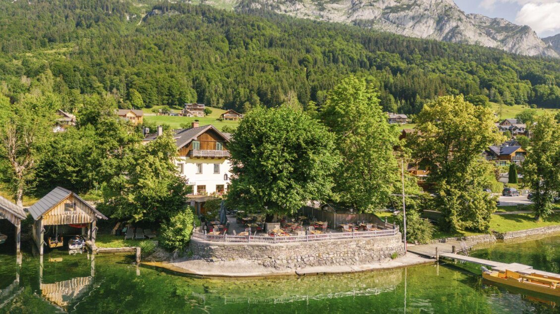 Eine idyllische Landschaft mit einem klaren See und umgeben von Bäumen. Im Hintergrund sind Berggipfel und traditionelle Häuser zu sehen. | © DraganTadic