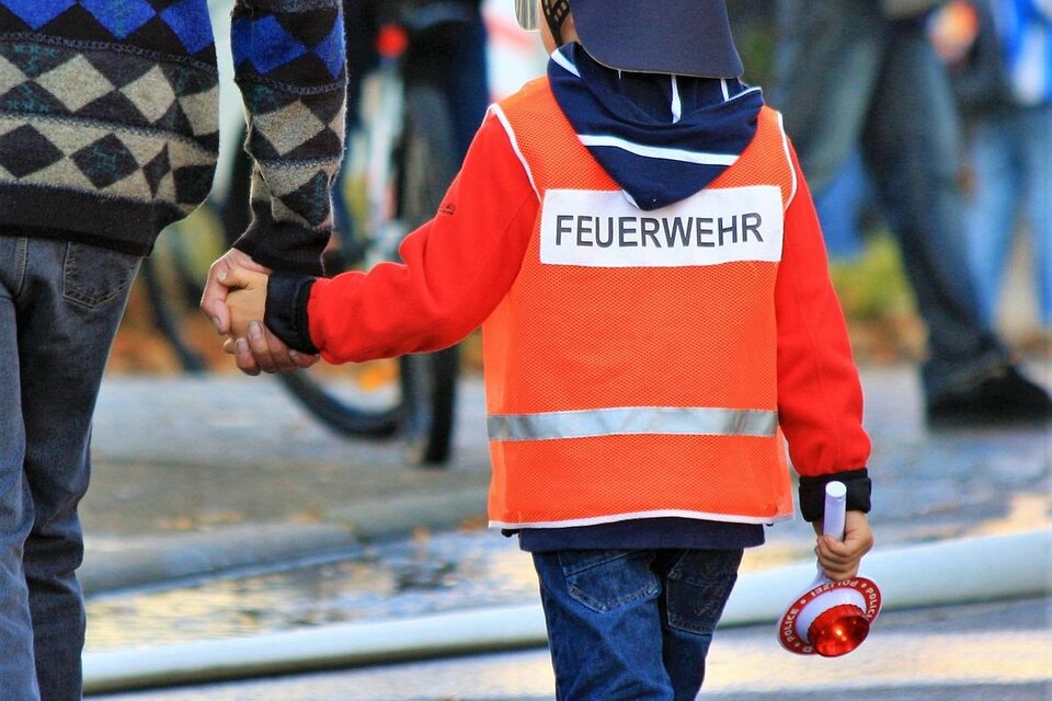 Ein Kind in einer Feuerwehrjacke hält die Hand eines Erwachsenen. Das Kind trägt einen Feuerwehrhelm und hat eine kleine Sirene in der anderen Hand.