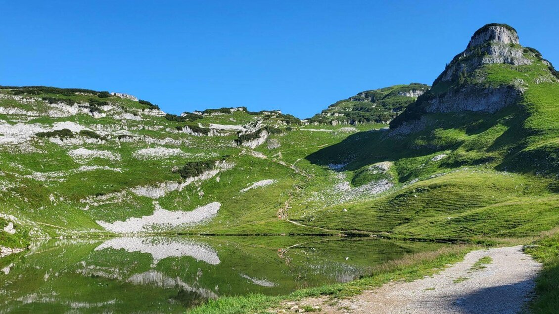 Eine grüne Berglandschaft mit einem klaren See und beeindruckenden Felsen. Der Himmel ist blau und strahlend. | © Petra Kirchschlager