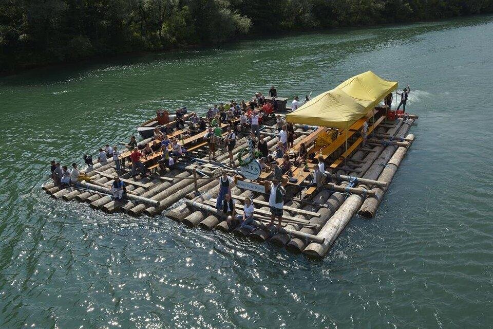 Eine große Floßfahrt mit vielen Menschen auf einem Fluss. Es gibt Bänke und ein gelbes Dach für Schatten. | © Floß & Co