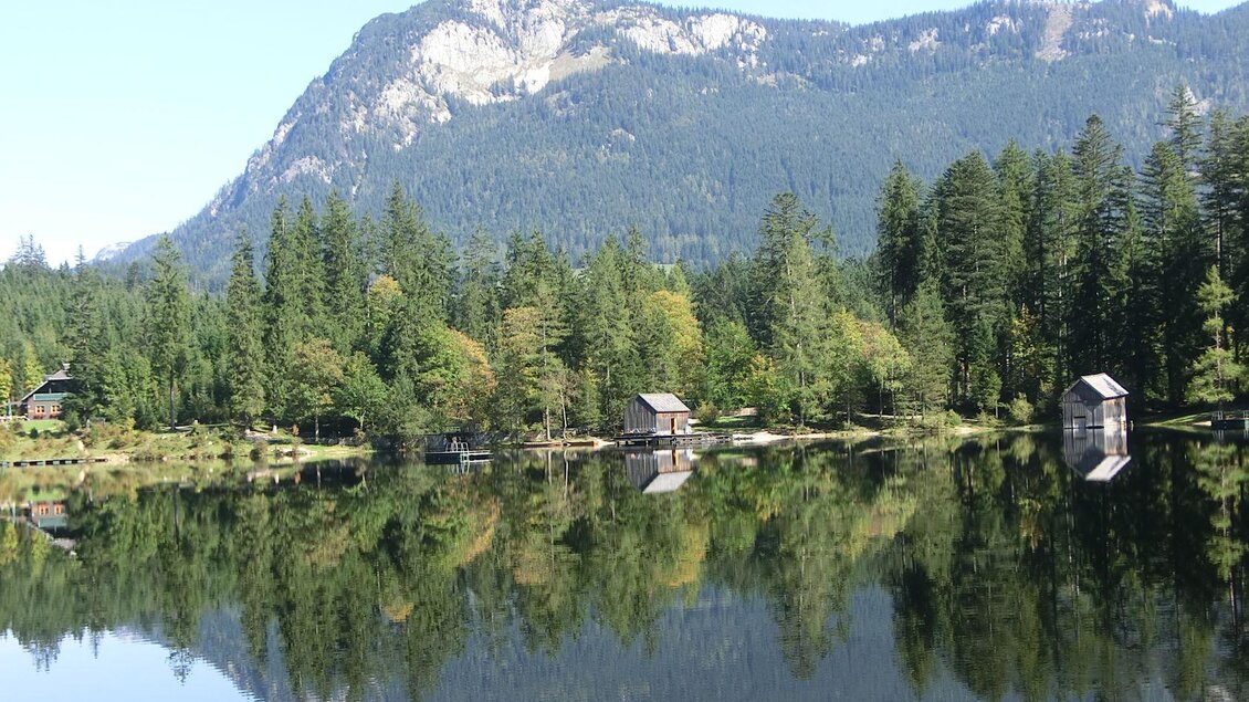 Ein ruhiger See umgeben von dichtem Wald und Bergen. Die Hütten spiegeln sich im klaren Wasser. | © TVB Ausseerland Salzkammergut-Tom Lamm