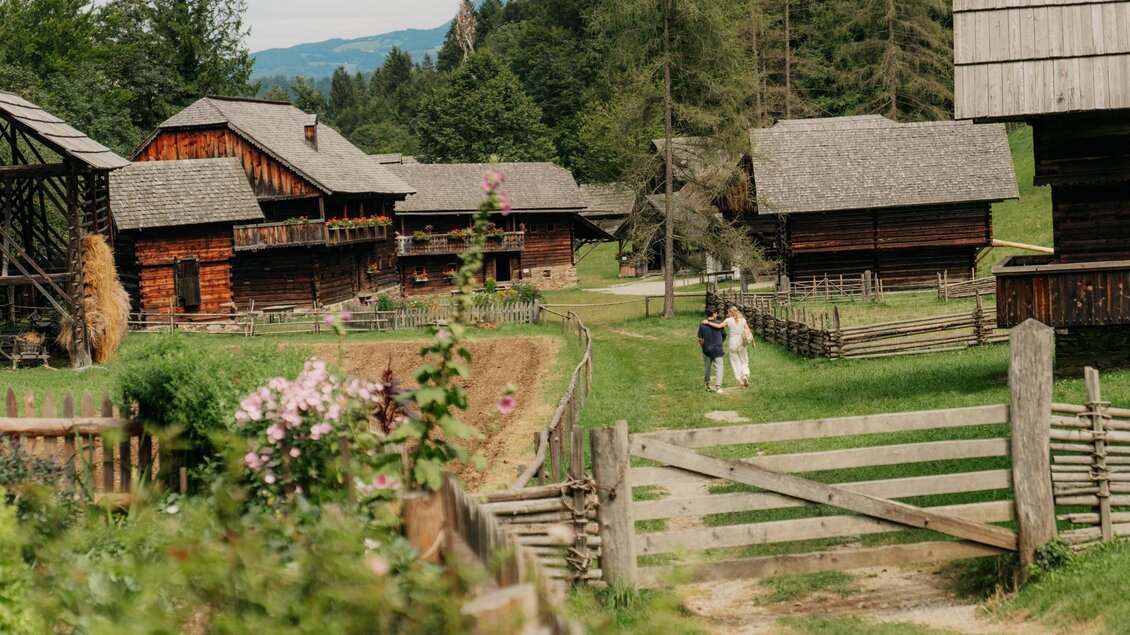Eine ländliche Szenerie mit traditionellen Holzhäusern und einer grünen Wiese. Im Hintergrund gehen zwei Personen gemütlich entlang des Weges. | © (c) Region Graz - studio draussen
