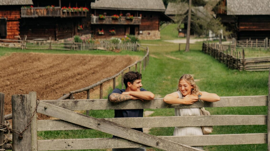 Zwei Personen lehnen am Holztor vor historischen Bauernhäusern im Freilichtmuseum Stübing. | © Region Graz-studio draussen