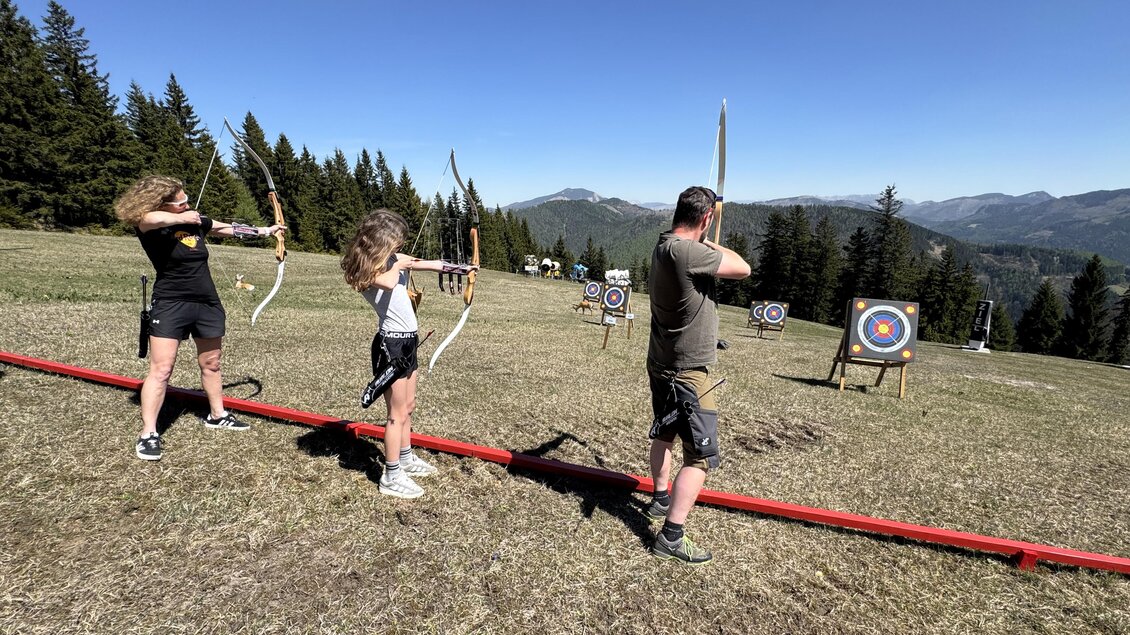 Drei Personen stehen im Freien und üben mit Bögen auf Zielscheiben. Im Hintergrund sind Bäume und Berge zu sehen. | © MariazellerBürgeralpe