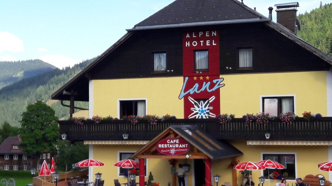 Ein charmantes Alpenhotel mit bunten Blumenfenstern. Vor dem Hotel befindet sich ein einladendes Café mit Schirmen und einer schönen Aussicht auf die Berge. | © Alpenhotel Lanz