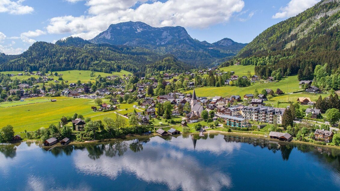 Eine malerische Landschaft mit einem klaren See, umgeben von grünen Wiesen und Bergen. Im Hintergrund ist ein kleines Dorf mit traditionellen Gebäuden zu sehen. | © Drohnenfotos.at-Andreas Tischler