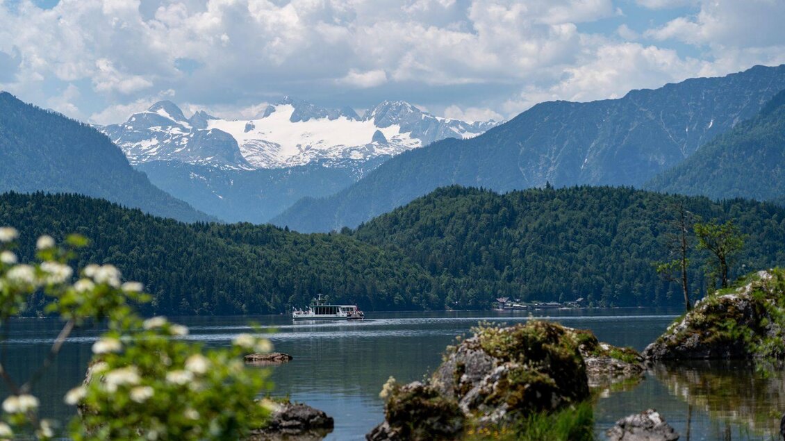 Ein ruhiger See umgeben von grünen Bergen und schneebedeckten Gipfeln im Hintergrund. Ein Schiff fährt auf dem Wasser und die Wolken sind am Himmel sichtbar. | © Salzkammergut-Altaussee_Oesterreich-Werbung_Cross-Media-Redaktion