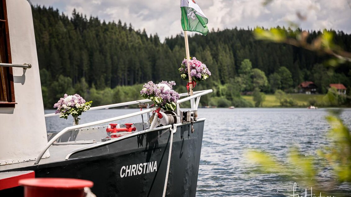 Ein Boot mit dem Namen "CHRISTINA" am Wasser, geschmückt mit Blumen. Im Hintergrund sind bewaldete Hügel und eine ruhige Landschaft zu sehen. | © www.mariazell.blog
