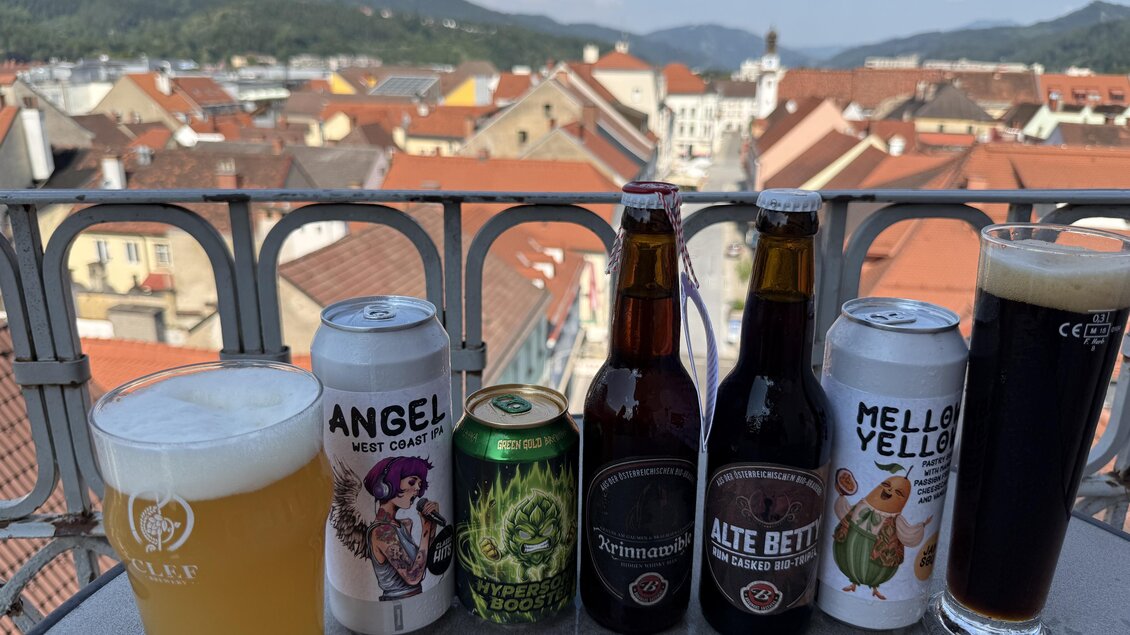 Eine Auswahl an verschiedenen Biersorten steht auf einem Tisch mit Blick auf eine Stadtlandschaft. Im Hintergrund sind Dächer und Berge unter einem blauen Himmel sichtbar. | © TVB Erzberg-Leoben