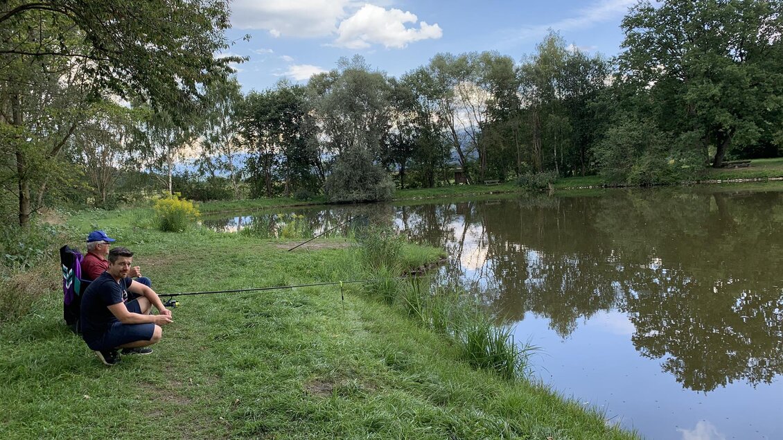 Drei Personen sitzen am Ufer eines ruhigen Teichs und angeln. Der Himmel ist teilweise bewölkt und die Natur um sie herum ist grün und einladend. | © Anita Fössl