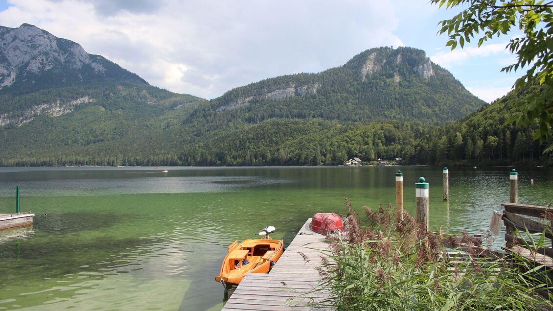 Ein ruhiger See umgeben von hohen Bergen und dichten Wäldern. Am Ufer liegt ein Holzsteg mit einem kleinen orangefarbenen Boot. | © Viola Lechner