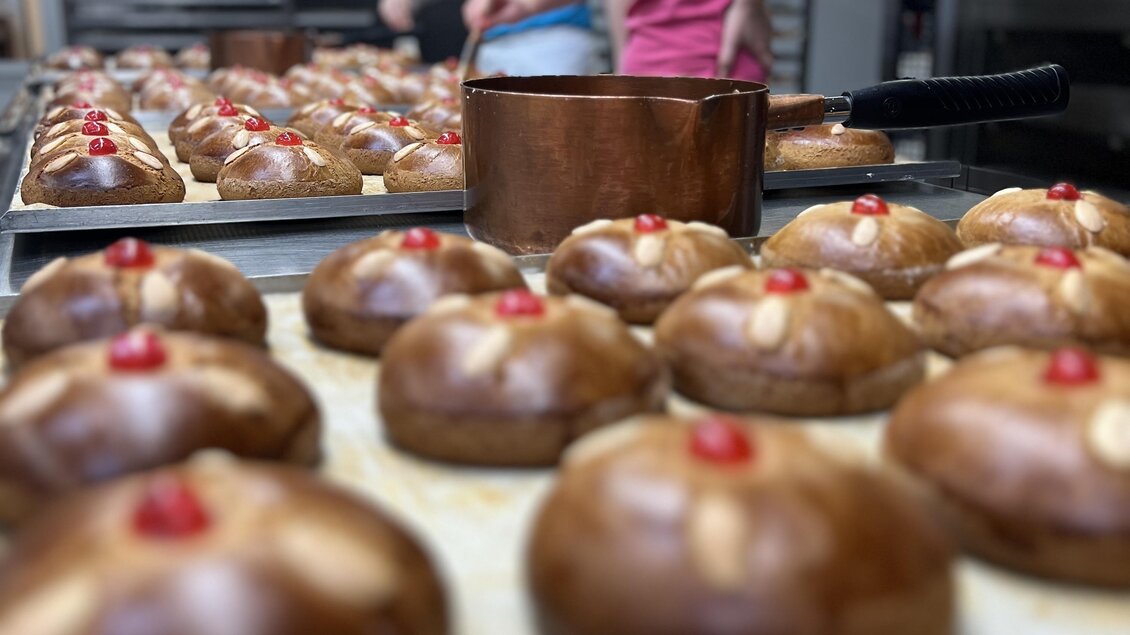 Zwei Bäckerinnen in der Backstube arbeiten an frischen Gebäckstücken. Auf dem Tisch sind viele glänzende Brötchen mit Kirschen dekoriert. | © Konditorei Regner