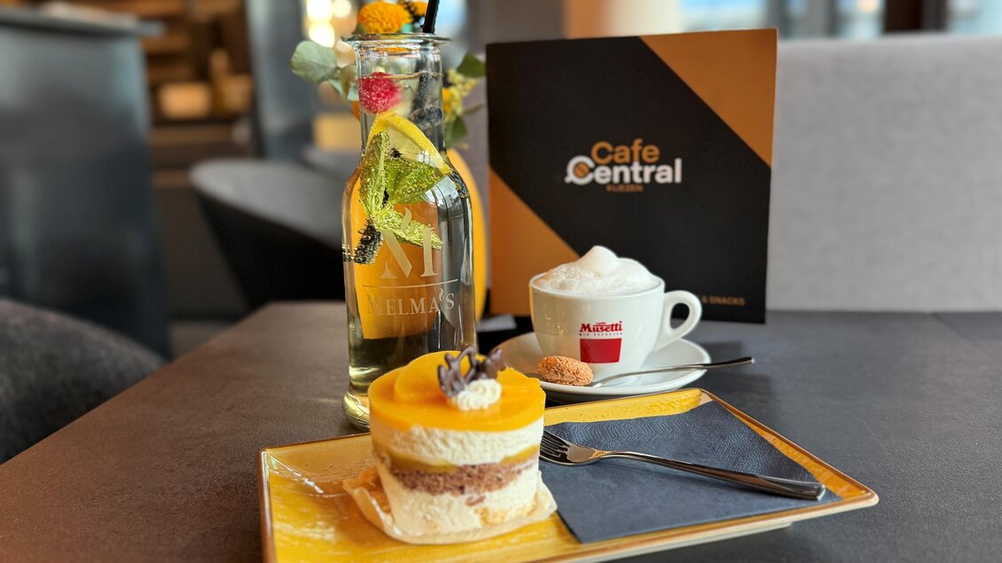 Ein leckerer Kuchen mit einer Mango-Deckschicht und einer Tasse Kaffee auf einem Tisch. Im Hintergrund steht eine Flasche und eine Speisekarte des Café Central. | © Cafe Central