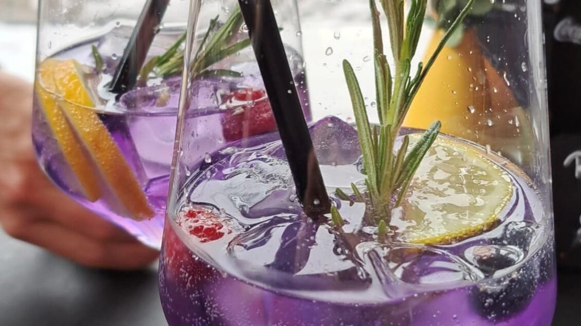 Zwei Gläser mit lila Getränken, verziert mit Zitronenscheiben, Beeren und Rosmarin. Im Hintergrund sind eine Hand und eine Blume zu sehen. | © Cafe Central