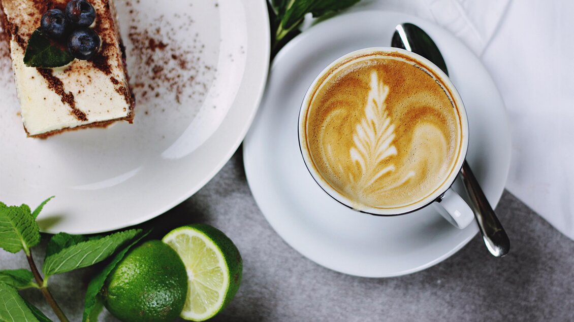 Ein schöner Kaffee mit kunstvollem Milchschaum und ein Stück Torte sind auf einem Tisch angerichtet. Frische Limetten und Minzblätter ergänzen das Bild. | © Pixabay