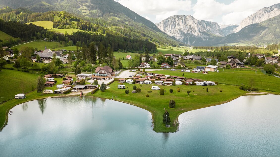Eine malerische Landschaft mit einem ruhigen See und sanften Hügeln. Im Hintergrund erheben sich majestätische Berge und idyllische Wohnmobile sind auf der Wiese verteilt. | © Hannes Zenz