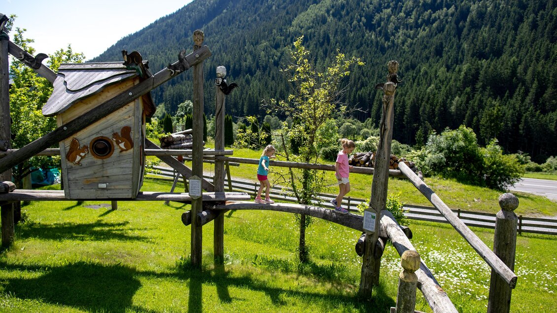 Ein Spielplatz in der Natur mit einer Holzstruktur und zwei Kindern, die darauf spielen. Im Hintergrund sind grüne Berge und Bäume zu sehen.