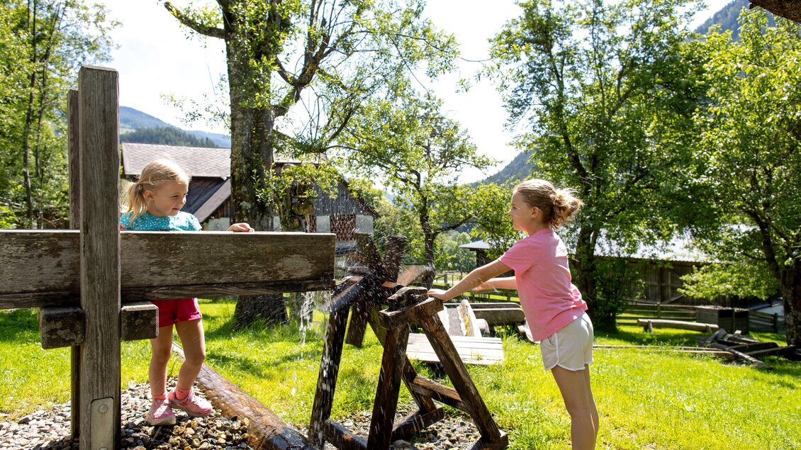 Zwei Mädchen spielen im Freien an einem schönen, sonnigen Tag. Sie stehen neben einem alten Holzschwengelpumpen und einer Holzstruktur.