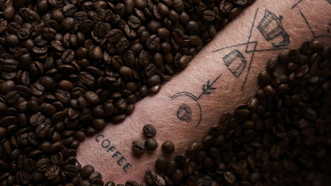 Ein Arm ist in Kaffeebohnen eingetaucht, mit verschiedenen Tattoos sichtbar. Das Wort „COFFEE“ ist deutlich zu lesen. | © Nationalpark Gesäuse