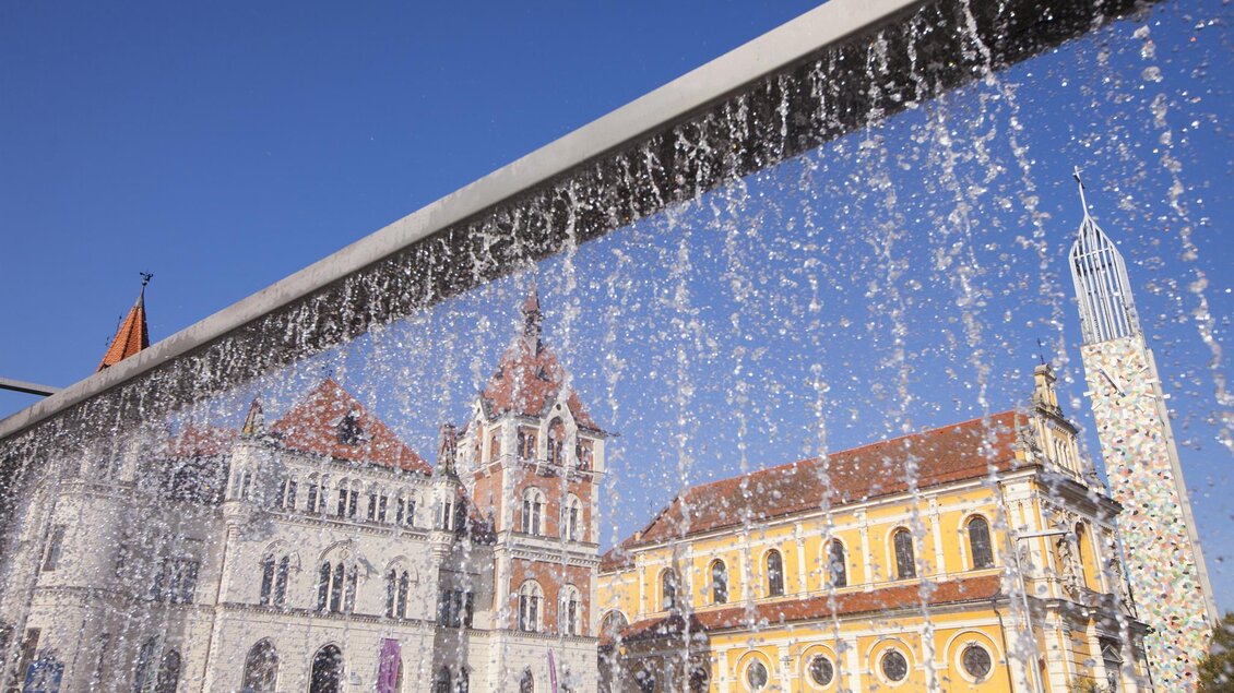 Ein modernes Wasserfall-Design vor historischen Gebäuden. Im Hintergrund sind zwei auffällige Gebäude und ein blauer Himmel sichtbar. | © Tourismusverband Feldbach/ B. Bergmann