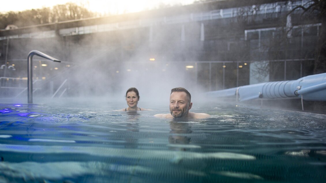 Zwei Personen entspannen in einem warmen, dampfenden Pool. Im Hintergrund sind Bäume und eine moderne Architektur zu sehen. | © Steiermark Tourismus