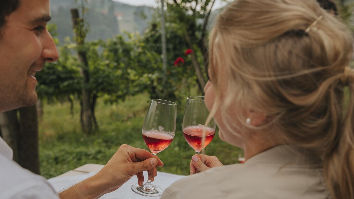Ein Paar sitzt in einem Weingarten und prostet sich mit Gläsern Rosé zu. Im Hintergrund sind grüne Reben und sanfte Hügel zu sehen. | © TV Südsteiermark - Lupi Spuma