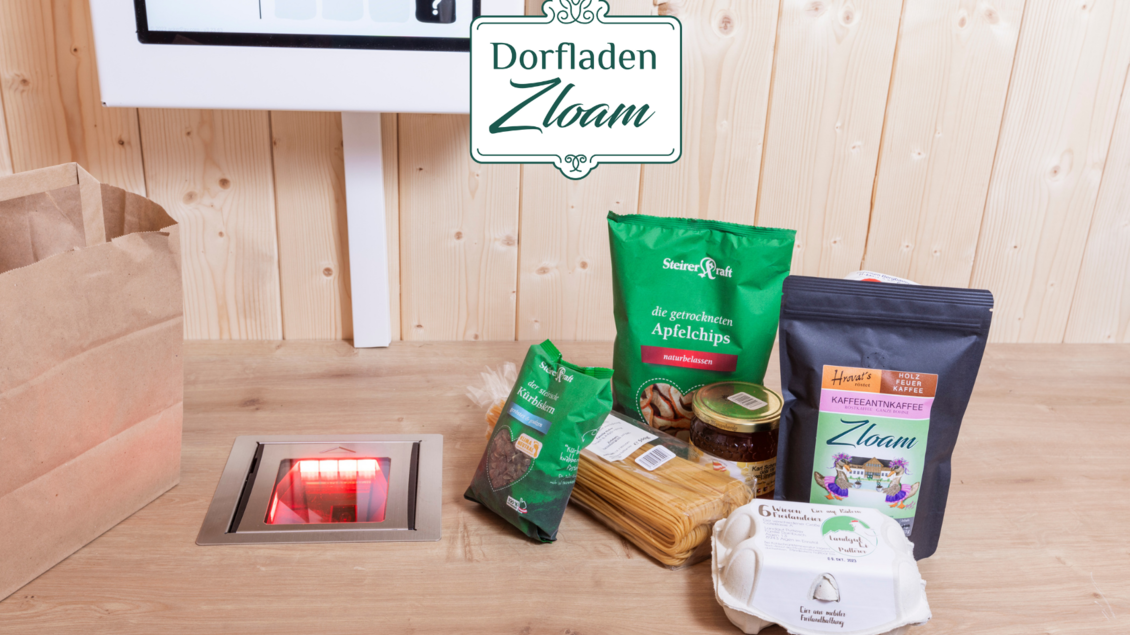 Ein Tisch mit verschiedenen Lebensmitteln ist zu sehen, darunter Nudeln, Äpfelchips, eine Konservendose und Eier. Im Hintergrund befindet sich ein Bildschirm mit dem Logo „Dorfladen Zloam“. | © Narzissendorf Zloam, www.zloam.at