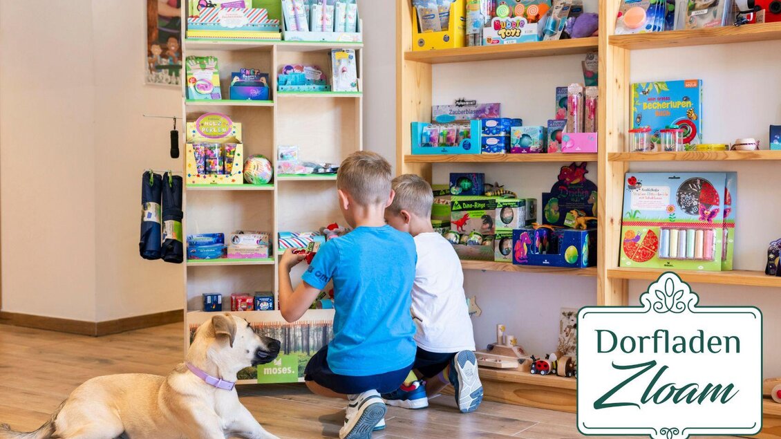 Zwei Kinder schauen sich Spielzeug in einem Dorfladen an. Ein Hund liegt entspannt auf dem Boden. | © www.zloam.at