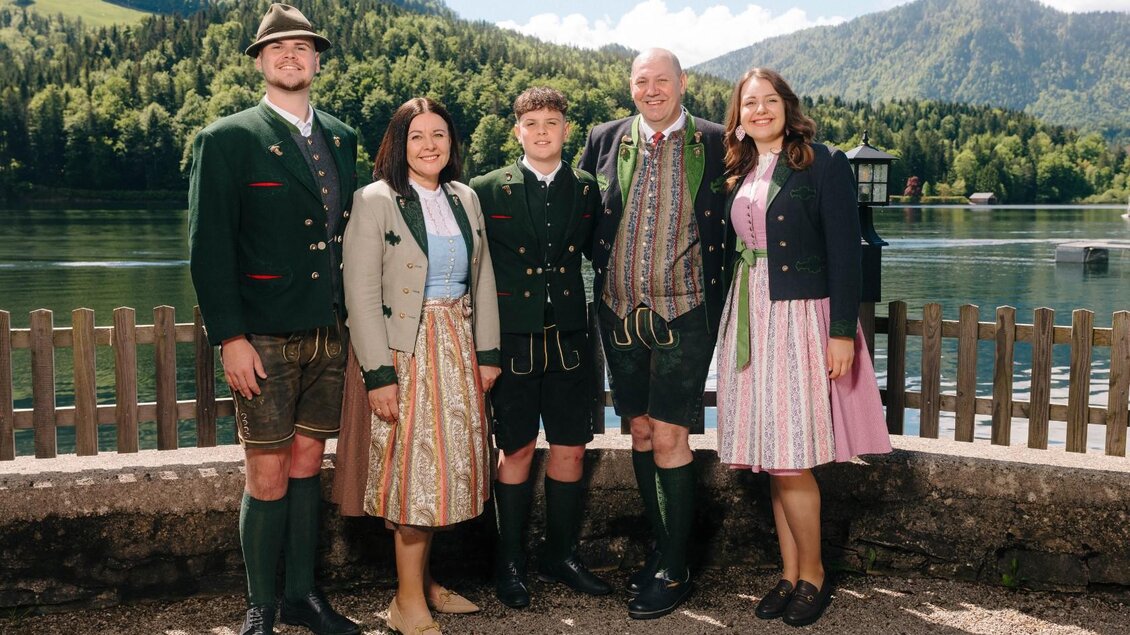 Eine Gruppe von fünf Personen in traditioneller Tracht steht am Ufer eines Sees. Im Hintergrund sind grüne Berge und ein klarer Himmel zu sehen. | © Gasthaus Stöckl